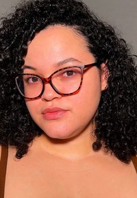 Skilpaddedekkbriller med vinklede rammer, fremhevet av glansfulle detaljer. Krøllete hår, lys hudtone og enkel brun topp.