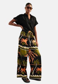 Camisa negra de manga corta combinada con pantalones de pierna ancha que presentan un vibrante patrón de palmeras y sol en naranja y verde sobre un fondo negro.
