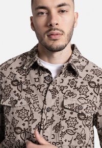 Πουκάμισο από κατσαρίνα με λουλουδάτο μοτίβο σε μπεζ χρώμα και καφέ λεπτομέρειες, διαθέτει κουμπιά μπροστά, δύο τσέπες στο στήθος και κλασικό γιακά.