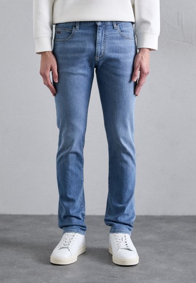 Jeans in denim azzurro chiaro con vestibilità slim, design a cinque tasche, dettagli di cucitura sottili e texture consumata, indossati con sneakers bianche.