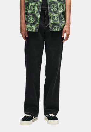 Persona che indossa pantaloni neri a gamba larga, sneakers nere con suole e lacci bianchi e una camicia verde a fantasia con simboli di pace.