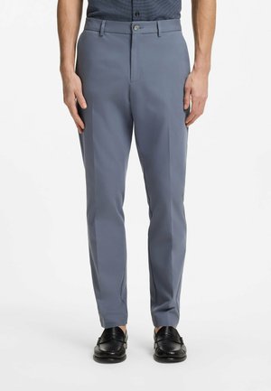 Uomo con pantaloni grigi a gamba dritta con tasche frontali e mocassini neri, in piedi su uno sfondo bianco.