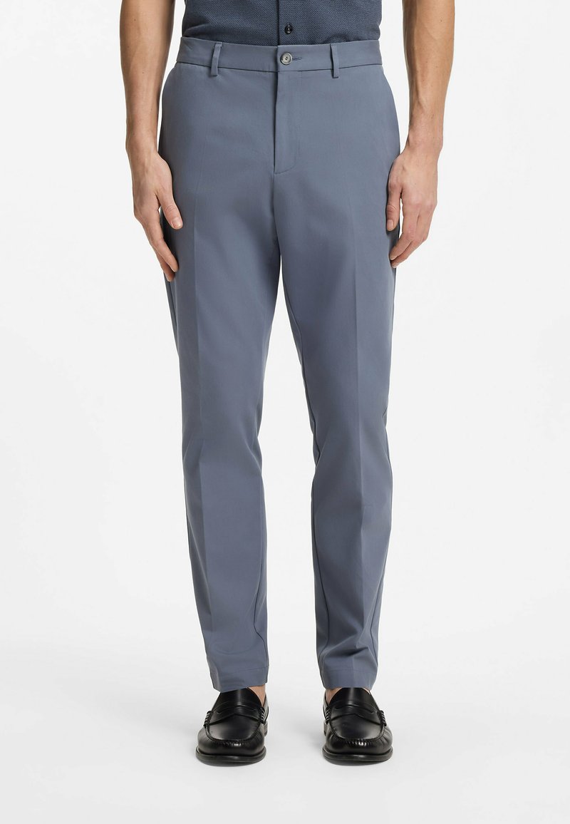 Homme portant un pantalon gris droit avec poches avant et des mocassins noirs, debout devant un fond blanc.