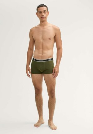 Männliches Model steht barfuß und trägt olivgrüne Boxershorts mit einem schwarzen Bund vor einem schlichten hellen Hintergrund.