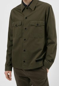 Chaqueta ligera de camisa en verde oliva, cierre frontal de botones, dos bolsillos en el pecho, botones negros y detalle de logo en el lateral. Textura de tela suave.