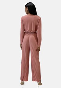 Tuta rosa a maniche lunghe con un design plissettato testurizzato e una cintura elastica. Presenta pantaloni larghi e sandali con tacco lucidi.