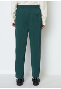 Grüne, elastische Hose mit glatter Textur. Verfügt über eine Gesäßtasche mit einem Knopf und ein gerades Bein-Design. Trägt schwarze Schuhe.