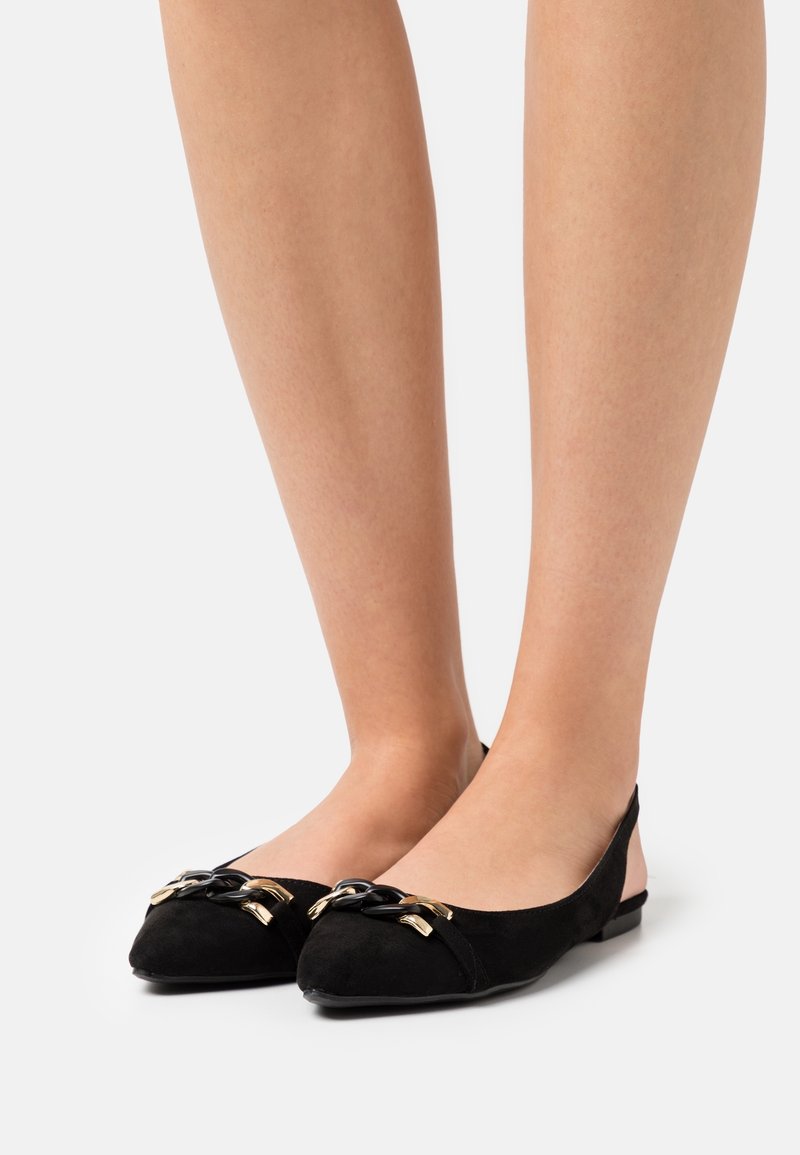 Jambes portant des ballerines pointues en daim noir avec un détail de chaîne noire et dorée sur les bouts, sur un fond neutre.