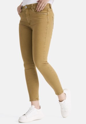 Beige Skinny Jeans aus Denim mit Reißverschluss, vorderen und hinteren Taschen sowie tailliertem Design, kombiniert mit weißen Sneakers.
