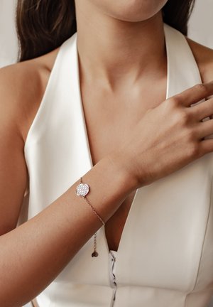 Frau in weißem ärmellosem Oberteil zeigt ein filigranes Armband aus Roségold mit rundem, gemustertem Anhänger am Handgelenk.