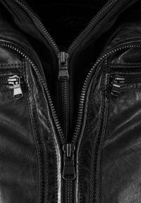 Nærbillede af en sort læderjakke med tre synlige sølv lynlåse, en centralt og to på brystlommerne, delvist åbnet.