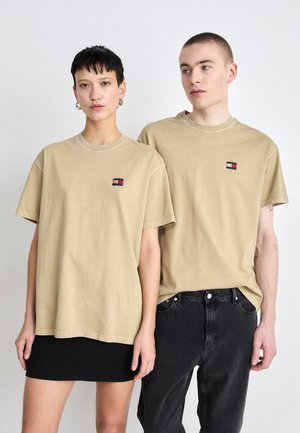 T-shirts en coton beige avec un petit patch multicolore, présentant des manches courtes et une coupe décontractée, portés avec une jupe noire et un jean foncé.