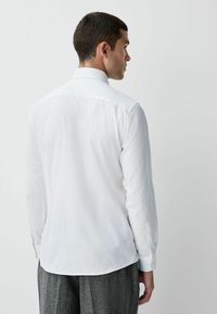 Valkoinen pitkähihaainen paita, jossa on levitetty kaulus, nappiriippuiset hihansuut ja sileä pinta. Suora helma, jossa sivureiät, istuva malli.