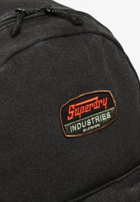 Czarny plecak z tkaniny o teksturowanej powierzchni, z wyhaftowanym logo "Superdry Industries" w kolorze czerwonym i zielonym oraz czarnym zamek błyskawiczny.