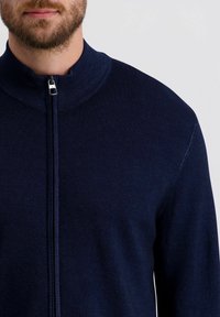 Marineblauer Reißverschluss-Pullover mit hohem Kragen, strukturiertem Rippdesign und silbernem Reißverschluss. Der Stoff wirkt weich und leicht. Keine sichtbaren Muster.