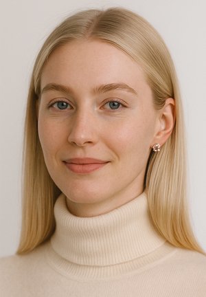 Närbild på en person som bär en krämfärgad turtleneck-tröja, med en blommig studörhänge. Slät hud, rakt blont hår och neutral uttryck.