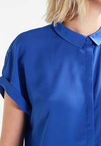 Blå blus med kort ärm, tillverkad av mjukt tyg. Den har en spetsig krage och knappstängning framtill, rullade ärmslut och en avslappnad passform.