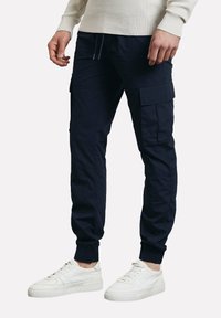 Pantalon cargo bleu marine avec plusieurs poches cargo, poignets élastiques et taille à cordon, fabriqué en tissu durable, assorti à des baskets blanches.