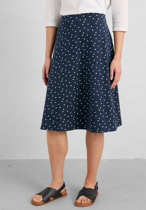 Femme portant une jupe bleu marine longueur genou à pois blancs, un top blanc à manches trois-quarts et des sandales noires avec des semelles beiges.