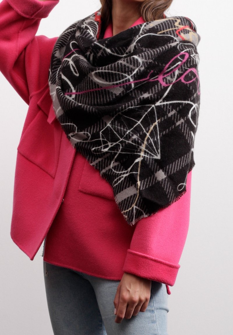 Femme portant un manteau rose vif et un jean bleu clair, enveloppée d'une écharpe noire à motifs avec des lignes abstraites et des accents de texte.