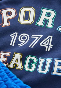 Morská modrá tkanina s výrazným, viacfarebným textom: "ŠPORTOVÁ LIGA" a "1974" v bielej, oranžovej, žltej a zelenej akcentoch. Textúrovaný modrý lem.