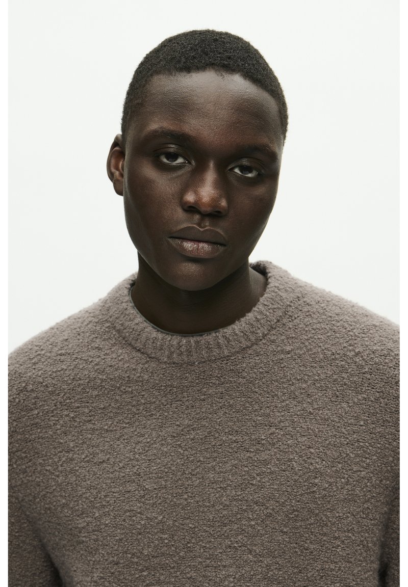 Un pull en maille marron texturé avec un col rond et un motif de tricot confortable, présentant une surface douce et légèrement pelucheuse. Vue en gros plan du vêtement.