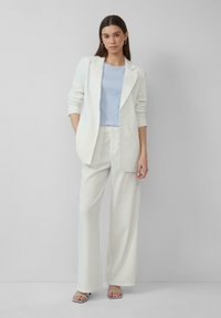 Un costume blanc léger à rayures, avec une veste ajustée et un pantalon large, associé à un haut bleu clair et des sandales argentées à lanières.