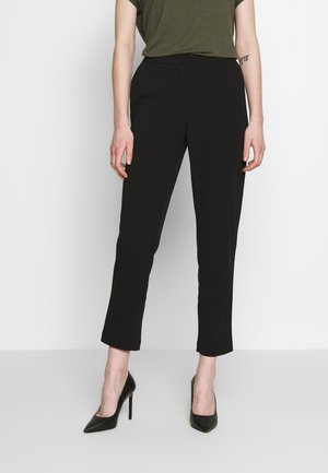 Person wearing black straight-leg ankle pants, olive green short-sleeve top, and black high-heeled shoes, standing against a white wall.