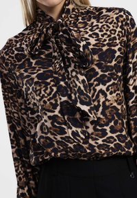 Blusa a stampa leopardata con scollatura annodata, texture setosa e una palette di colori marrone e crema, abbinata a pantaloni neri sartoriali.
