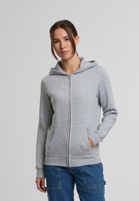Felpa grigia con zip realizzata in materiale morbido, dotata di cappuccio, tasche frontali e polsini a costine. Abbinata a pantaloni in denim blu.