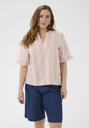 Femme aux longs cheveux bruns portant un chemisier rose à rayures avec des manches volantées et un short en jean bleu foncé arrivant aux genoux.