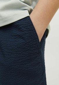 Shorts texturés en bleu marine avec un motif en relief et des poches latérales, portés avec une chemise gris clair, présentant une ourlet visible et un tissu lisse.