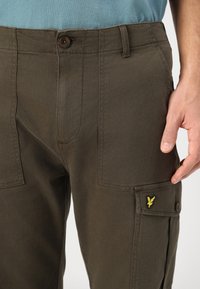 Tmavo zelené cargo nohavice vyrobené z odolnej látky, s uzatváraním na gombík a bočnými vreckami. Výrazná žltá emblema blízko pravého vrecka.