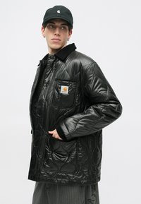 Czarna pikowana kurtka z kołnierzykiem, z przednią kieszenią z naszywką z logo. Noszona na ciemną, teksturowaną koszulę i szare, w paski spodnie.