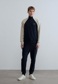 Μαύρο μπουφάν με φερμουάρ και εκρού μανίκια, συνδυασμένο με σκουρόχρωμα αθλητικά παντελόνια. Ομαλή υφή, μινιμαλιστικός σχεδιασμός, με λογότυπο στο στήθος και αθλητικά παπούτσια.