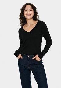 Görbe, sötét hajú nő mosolyog, fekete, hosszú ujjú V nyakú felsőt és sötétkék farmert visel, kezei a mellső zsebekben vannak.