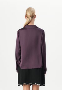 Blonďatá žena s nízkým culíkem má na sobě dlouhý purpurový top a černou sukni s stříbrným volánkovým lemem, viděná zezadu.