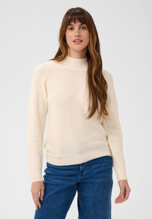 Femme aux longs cheveux bruns portant un pull crème et un jean bleu, debout devant un fond clair uni.