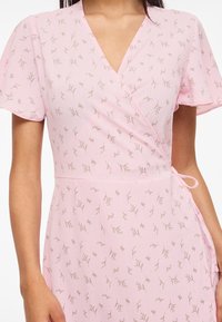 Ροζ φόρεμα τύπου κιμονό με κοντά μανίκια, λεπτό καφέ φλοράλ σχέδιο και λεπτομέρεια με κορδόνι στο πλάι στο σώμα μιας γυναίκας.