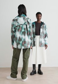 Two models are wearing a patterned, lightweight rain jacket in teal and brown, paired with olive green pants and a white skirt.