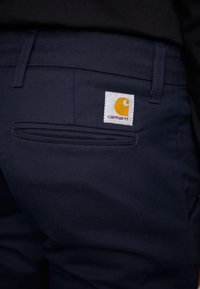 Pantalons bleu marine au design ajusté, avec une poche arrière et une étiquette en tissu blanc avec un logo orange. Texture lisse, style décontracté.