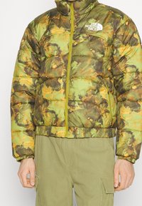 Prošívaná bunda v zeleném maskáčovém vzoru s oranžovými akcenty. Má vysoký límec, zapínání na zip a elastické manžety. Viditelné logo značky.