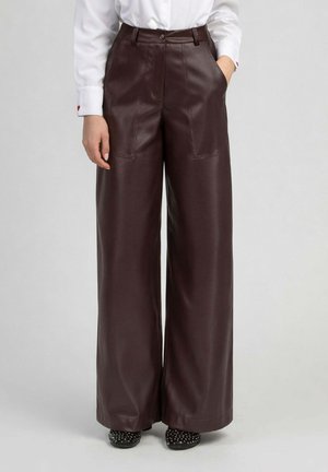Femme portant un pantalon en cuir marron foncé, taille haute et jambes larges, une chemise blanche et des chaussures noires à pois, debout avec une main dans la poche.