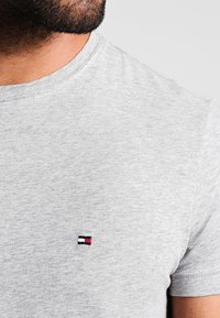 Graues Baumwoll-T-Shirt mit Rundhalsausschnitt, mit einem kleinen gestickten Logo in Rot, Weiß und Blau auf der linken Brust. Weicher Stoff.