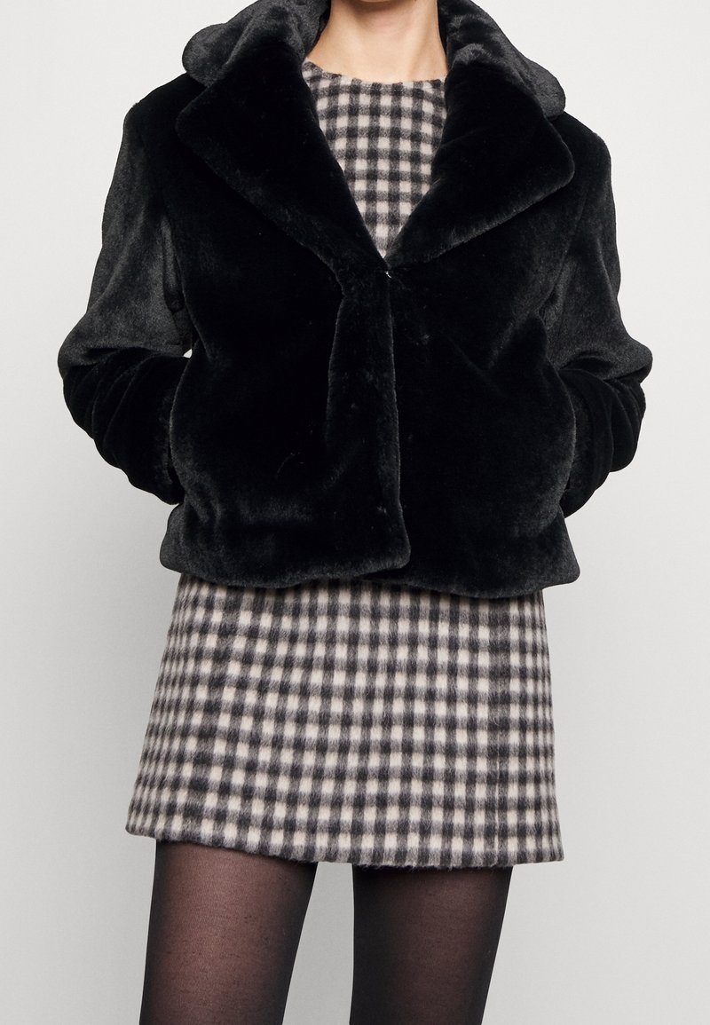 Veste en fausse fourrure noire avec large col portée sur une robe à carreaux noir et blanc et des collants noirs transparents.