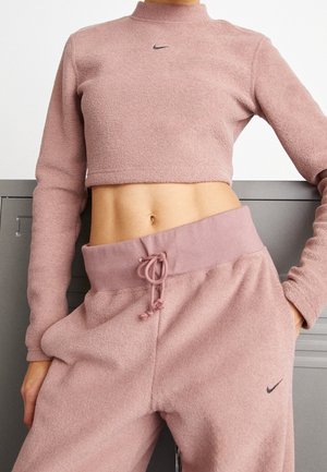 Kobieta ubrana w różowy krótki polarowy sweter Nike i pasujące spodnie dresowe ze ściągaczem w talii, stojąca z jedną ręką w kieszeni.