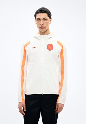 Młody mężczyzna zwrócony przodem, ubrany w białą kurtkę zapinaną na zamek z pomarańczowymi paskami na rękawach, z naszywką lwa i logo Nike.
