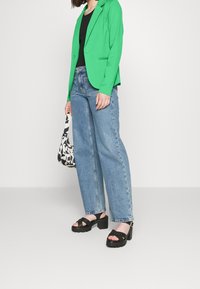 Zielona marynarka, czarny top, niebieskie jeansy z wysokim stanem oraz czarne sandały na platformie. Akcesoria to czarno-biała torebka z wzorem.