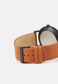 Brun läderarmband med en slät textur, utrustad med en svart metalklämma och en minimalistisk svart urtavla. Märkt med "SKAGEN".
