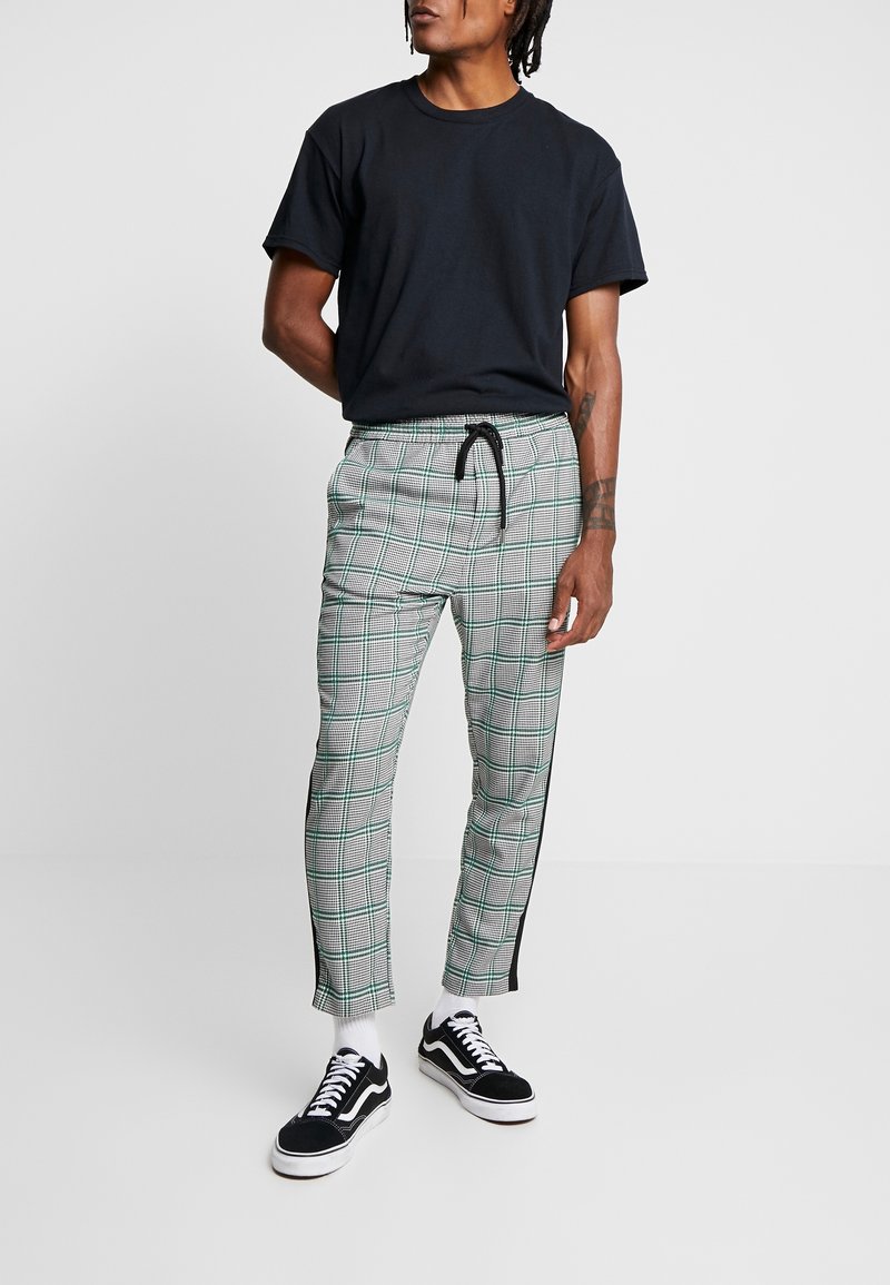 Homme portant un t-shirt noir, un pantalon gris à carreaux avec des bandes latérales noires, des chaussettes blanches et des baskets noir et blanc, debout avec une main derrière le dos.